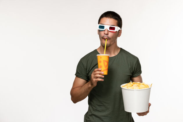视图正面图身穿绿色t恤的年轻男性手持马铃薯cips苏打水戴着d太阳镜在白墙上拍摄电影《孤独的电影》电影电影肖像