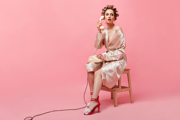 快乐穿着丝绸长袍 头上戴着卷发器的女人正坐在椅子上 在粉红色的墙上讲电话人电话脸
