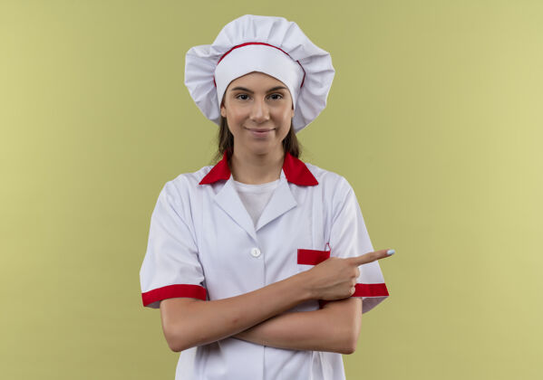 烹饪身着厨师制服的年轻自信的白人厨师女孩交叉双臂 指着旁边的绿色空间壁板年轻手臂