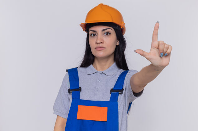 严重身穿施工制服 头戴安全帽的年轻建筑工人站在白色的墙上 严肃的脸上露出食指警告脸女人建筑