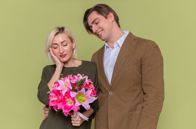情侣年轻漂亮的夫妇幸福的男人看着他可爱的女朋友和花束庆祝国际妇女节站在绿色的墙上爱花束快乐