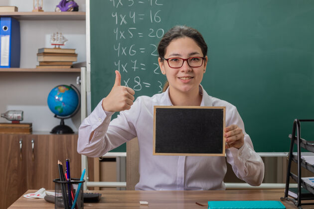 年轻人年轻的女老师戴着眼镜拿着小黑板讲课看着前面微笑着自信地竖起大拇指坐在教室黑板前的课桌上学校自信小