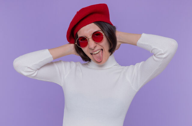 贝雷帽戴着贝雷帽 戴着红太阳镜 留着短发的年轻女子站在蓝色的墙壁上 快乐而快乐地伸出舌头望着前方年轻快乐太阳镜
