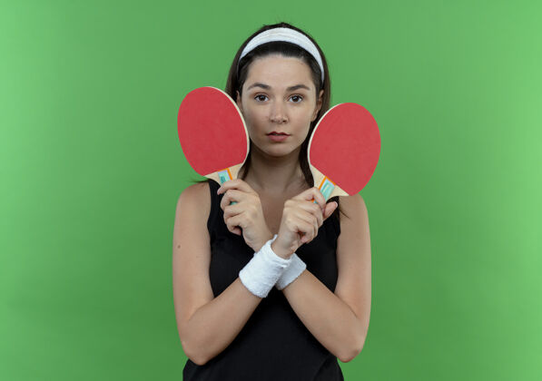 年轻戴着头巾拿着球拍打乒乓球的年轻健身女士 严肃的脸交叉着双手站在绿色的墙上脸桌子抱