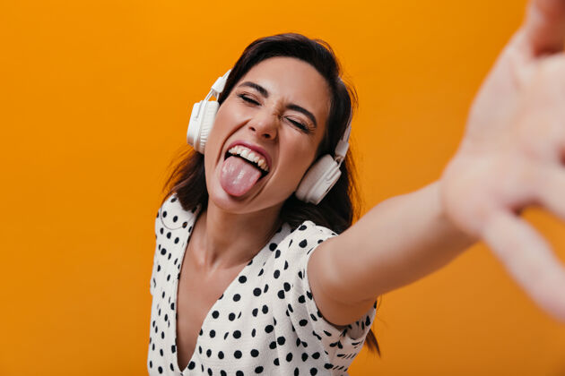 歌曲戴着耳机的女人露出舌头 在橙色背景上自拍开朗的女孩喜欢白色圆点衬衫圆点情绪休闲