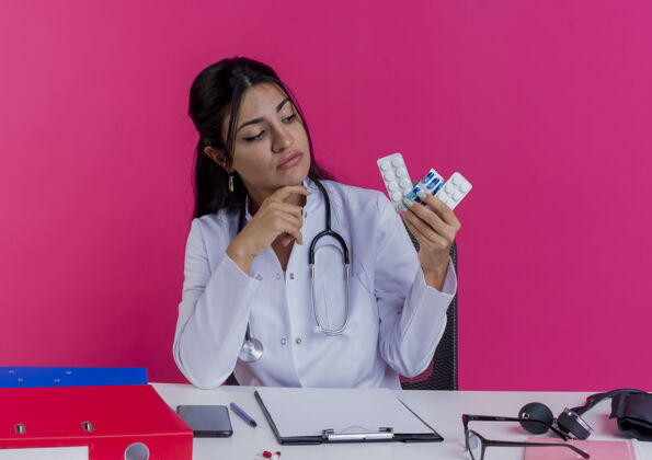 工具体贴的年轻女医生穿着医用长袍 手持听诊器坐在办公桌旁 手里拿着医疗工具 一边看着贴在粉红色墙上下巴的药品医疗触摸药物