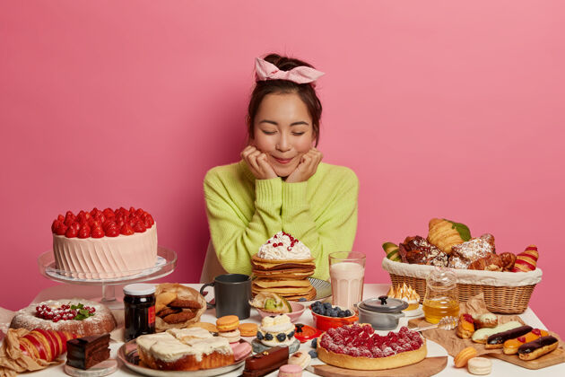 韩国人高兴的亚洲女人戴着头带和绿色套头衫 抱着下巴 胃口好 吃甜食 水果蛋糕 来参加生日聚会 孤立在粉红色的墙上饮食牛奶营养