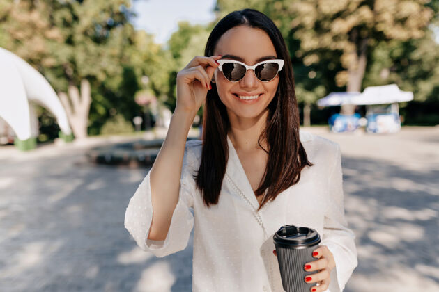 快乐在阳光明媚的城市公园里 穿着白衬衫 戴着白眼镜 在外面喝咖啡的快乐微笑的女人嘴唇魅力自信