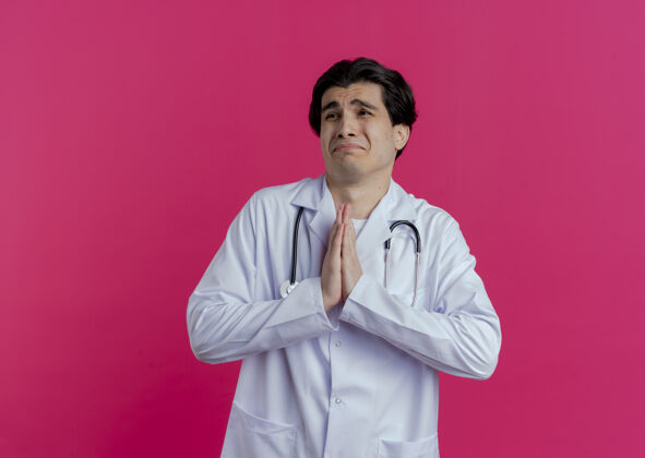 听诊器悲伤的年轻男医生穿着医用长袍和听诊器看着一边双手合十乞求隔离在粉红色的墙上有复印空间医生年轻手