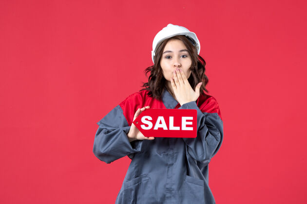 手势身穿制服 戴着安全帽的女工在隔离的红墙上展示销售图标和亲吻手势的正面近景肖像衬衫制服