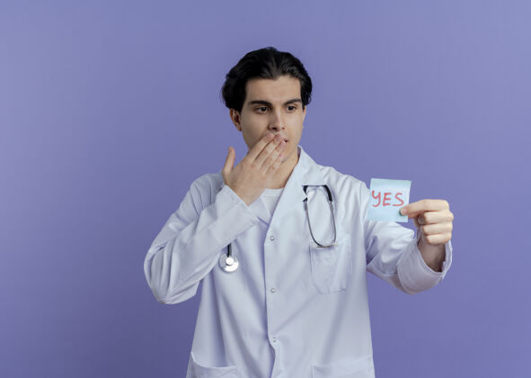 医学令人印象深刻的年轻男性医生穿着医用长袍和听诊器显示是的注意事项看着它保持手放在嘴上紫色墙上的复制空间隔离手长袍年轻