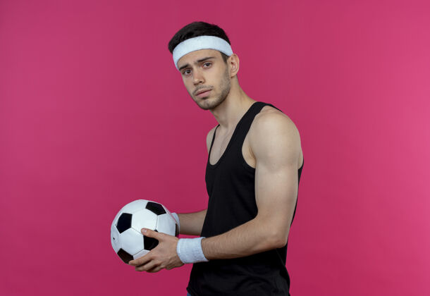 看着戴着头巾 表情严肃地拿着足球站在粉色墙壁上的年轻运动型男子表情头带运动
