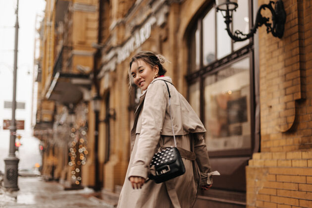亚洲年轻的亚洲女人兴高采烈地穿着时髦的风衣和黑色小包在城市里走来走去年轻城市背包