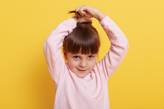 独自可爱的女孩做马尾辫 举手 看着镜头 穿着粉色休闲毛衣 孤立地站在黄色背景下 可爱的小女孩儿做发型肖像欢呼辫子