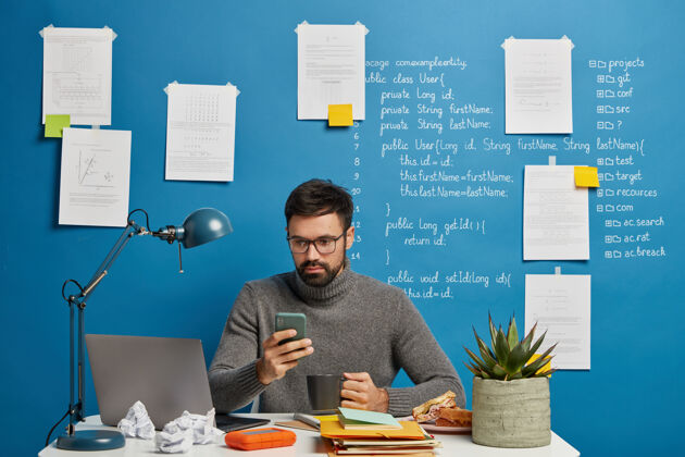 小工具It专业人员负责启动项目 更新手机上的软件和数据库 喝热饮料 坐在蓝色墙壁旁的桌面上写着信息笔记本电脑男手机
