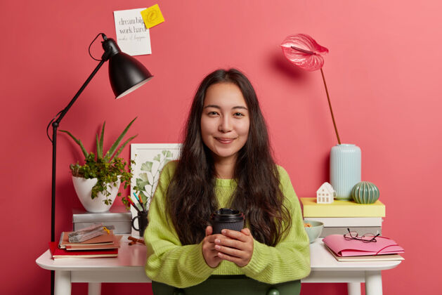 记事本漂亮的韩国女学者穿着绿色毛衣 拿着外卖咖啡杯 在家里准备研究 拿着鲜花对着舒适的工作场所摆姿势粘的笔记房子