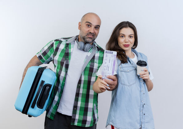 旅行高兴的成人旅行者夫妇男子戴着耳机脖子上拿着手提箱 女子拿着塑料咖啡杯 两人都拿着车票看抱着塑料女人