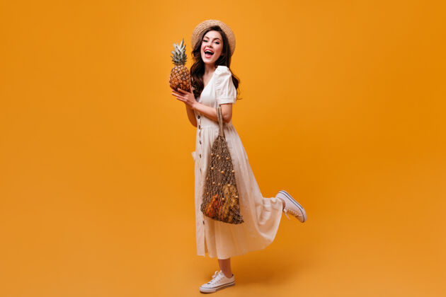 女性穿着迷笛裙 帽子和运动鞋的女人拿着购物袋和橘黄色背景上的菠萝年轻人脸头发