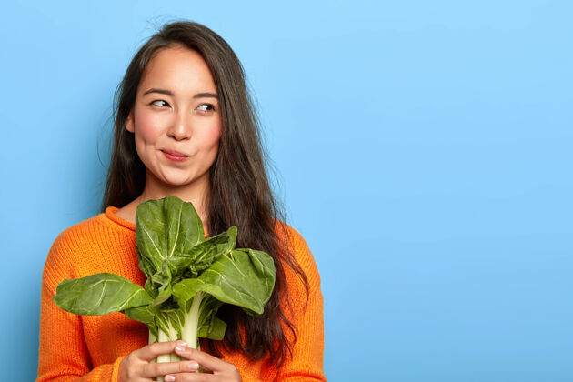 沙拉漂亮的年轻女子拿着新鲜的绿色蔬菜 在家里吃健康食品 用食品做素菜沙拉 穿着橙色套头衫 在室内摆姿势满意沉思套头衫