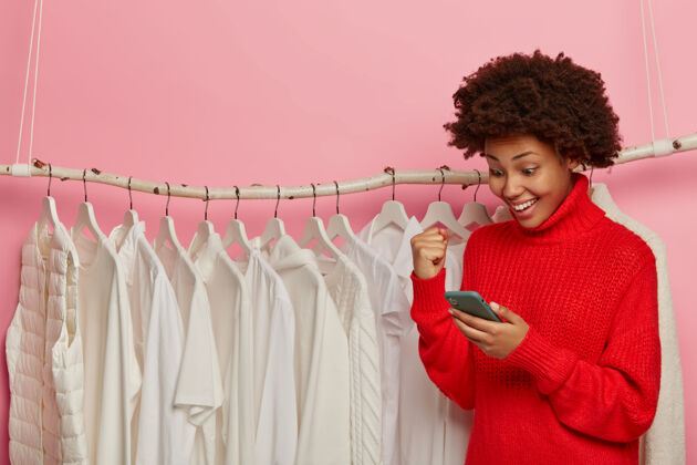 年轻人美国黑人妇女握紧拳头欢呼 积极地看着智能手机 对着衣架上的白色衣服摆姿势 在精品店购物欢呼个人资料购物