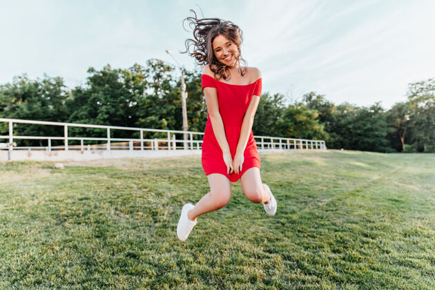 光激发了年轻女子在公园里跳跃和微笑的灵感穿着红裙子的漂亮黑发女孩在夏天的周末玩得很开心花园感性户外