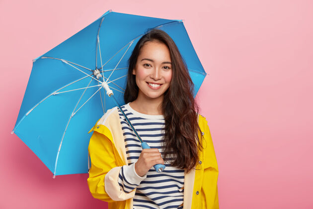 年轻喜气洋洋的黑发女人 黑色长发 穿着条纹套头衫 黄色雨衣 打着蓝色雨伞 在雨天散步条纹雨民族