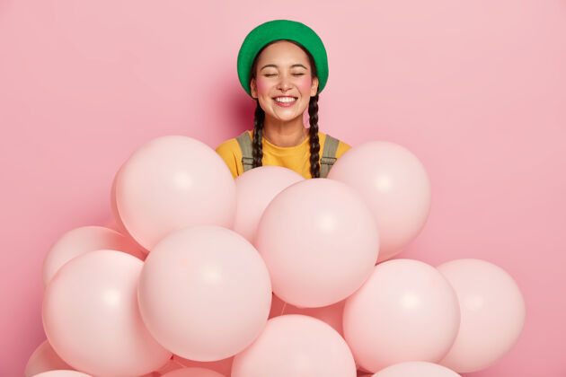 帽子戴绿帽的快乐亚洲女士的肖像 有两个辫子 胭脂脸颊 表达积极的情绪站在许多单色气球旁边节日休闲生日