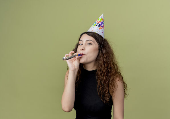 轻年轻漂亮的女人卷发在节日帽子吹哨子快乐和积极的生日派对概念站在轻墙帽子积极女人