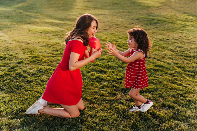 年轻穿着红色连衣裙的有趣的小孩和妈妈一起享受夏日美丽的黑发女人和女儿在草地上玩耍的户外照片积极黑发游戏