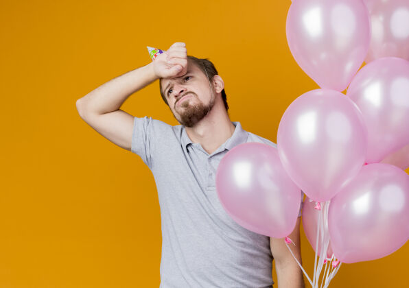 气球戴着节日帽的年轻人举着一堆气球站在橙色的墙上庆祝生日束男人看