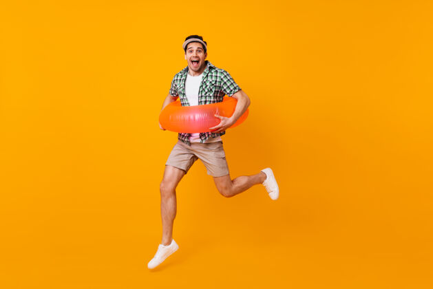 衣服棕色眼睛的深色男人穿着米色短裤和绿色t恤 在橙色的空间里用充气圈跳跃鸡尾酒水果行李