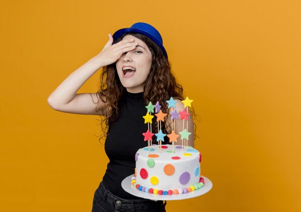 看着一个年轻漂亮的女人 卷发 戴着节日礼帽 手里拿着生日蛋糕 站在橙色的墙上 看起来既惊奇又快乐卷发年轻帽子