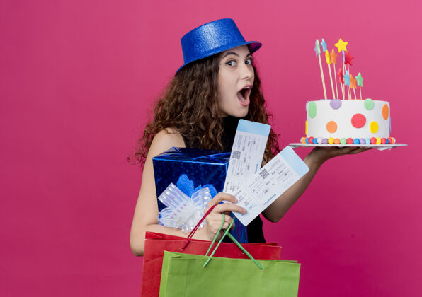 女人一位年轻漂亮的女士 卷发 戴着节日礼帽 手里拿着生日蛋糕礼盒和机票 站在粉色的墙上 快乐而兴奋的生日派对概念卷发抱着帽子