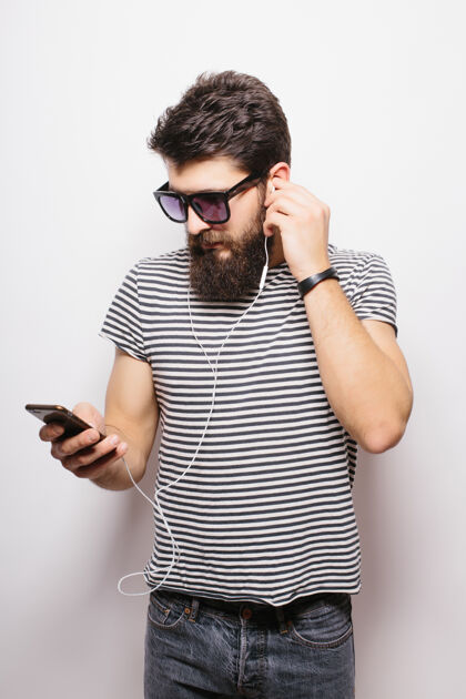 男性一个留着胡子的帅哥的肖像 戴着耳机 把手机孤立地放在白墙上表情年轻胡须