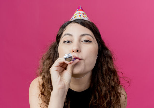 女人年轻漂亮的女人卷发在节日帽子吹口哨生日聚会的概念站在粉红色的墙上帽子卷发吹