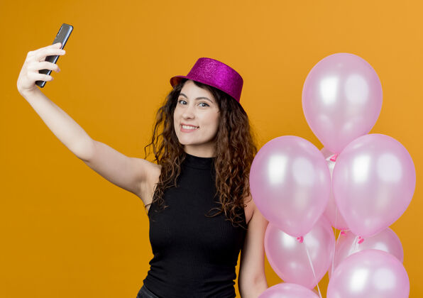 抱着一个年轻漂亮的女人 卷发 戴着节日礼帽 手里拿着一堆气球 站在橘色的墙上 自拍 开心地微笑着生日派对的概念空气自拍气球