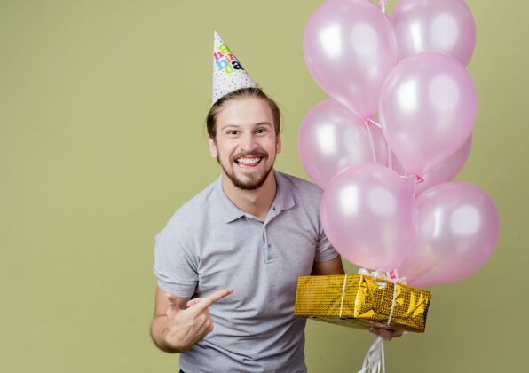 年轻戴着节日帽的年轻人举着生日礼物和气球庆祝生日 站在轻墙上 他高兴而兴奋地微笑着欢呼兴奋庆祝