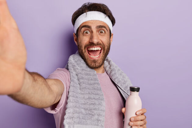 白种人快乐的运动员在训练中伸出手自拍 拿着水瓶 保持水分和健康 脖子上戴着毛巾 隔离在紫色的墙上紫色未刮胡子健康