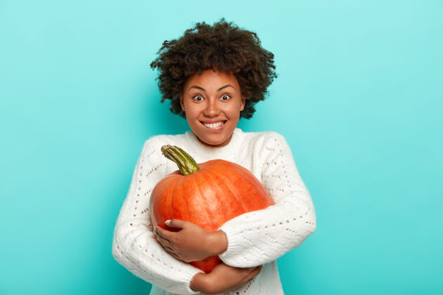 一个微笑着的女人 留着非洲式发型 咬着嘴唇 拥抱着橙色的大南瓜 秋天穿着白色的毛衣 在蓝色的背景下与世隔绝欢呼橙色摘
