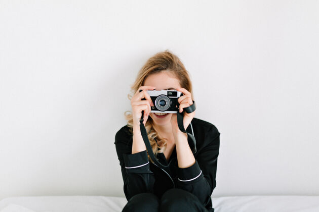 时尚金发的年轻女模特穿着黑色睡衣坐在白色的墙上 用复古相机拍照 放文字黑发皮肤欢呼