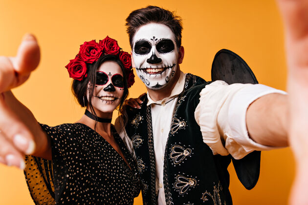 骷髅万圣节前夕 一个穿着睡衣的开朗男人和女朋友自拍橙色墙上的僵尸化妆的滑稽家伙死亡之日墨西哥化妆