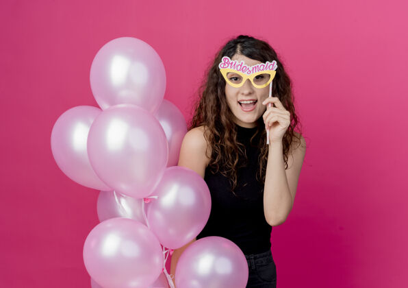 站着一个年轻漂亮的女人 卷发 手里拿着一束气球和一副纸眼镜 站在粉红色的墙上 开心地笑着女人气球束着