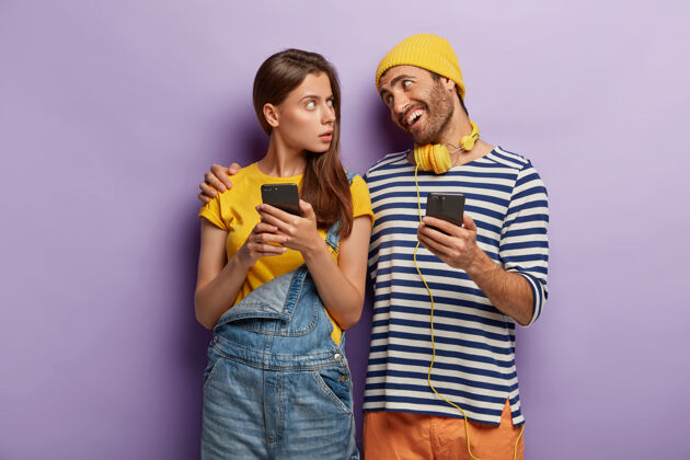 耳机快乐的男人拥抱忧心忡忡的女友 心平气和地交谈 使用现代手机 紧靠着紫色的墙壁站着拥抱条纹黑发