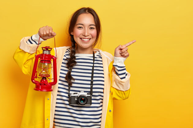 欢呼微笑的韩国女游客的半身镜头用食指指着远方 手持煤气灯 脖子上戴着复古相机 穿着随意 微笑愉快 在黄色背景上一览无余雨衣年轻复制空间