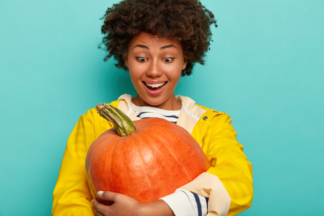 女孩快乐混血女拥抱大南瓜 穿着黄色雨衣 高兴地在自家花园里种大蔬菜 蓝色背景下的模特秋天的装饰抱着拥抱橙色
