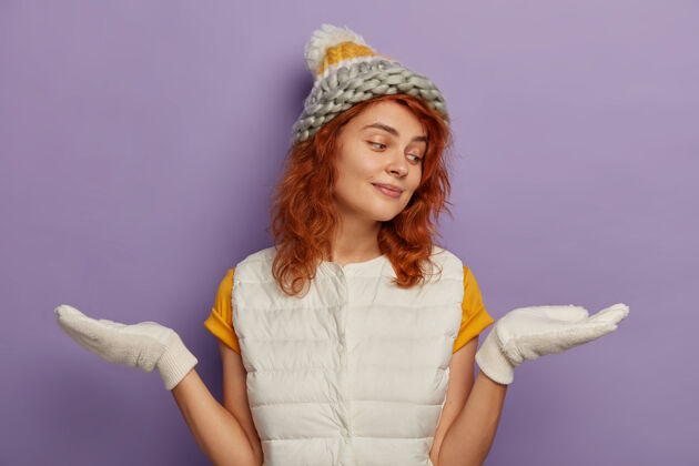 天气照片中的千禧一代女孩手心横着站着 假装拿着什么东西 在两件物品中选择 戴着帽子 戴着白手套 享受着寒假 在紫色的背景下与世隔绝舒适喜悦站立