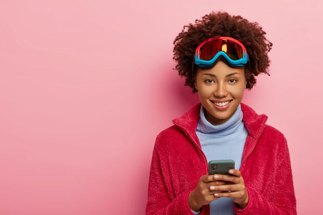设备室内拍摄的漂亮滑雪者女人使用智能手机设备 戴着滑雪面罩 对着镜头开心地微笑 隔着粉色的工作室墙美国手牙关
