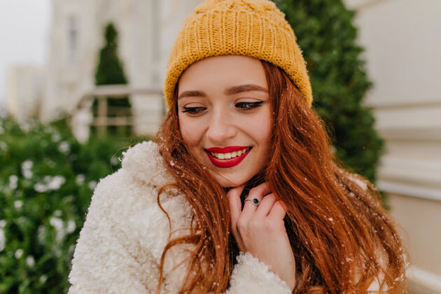 季节站在冷杉附近的迷人姜女的特写照片戴着冬天帽子的快乐微笑的女孩的户外照片时尚乐观欧洲