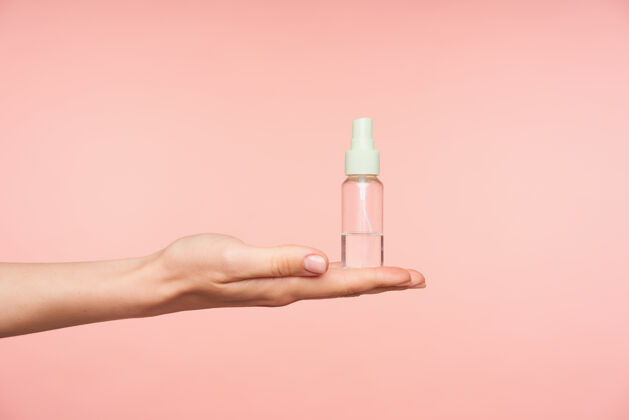 手臂侧视图精心打扮的女性的手保持手掌向上 同时举行透明喷雾瓶与液体对它 隔离在粉红色的背景举起皮肤白皙水平