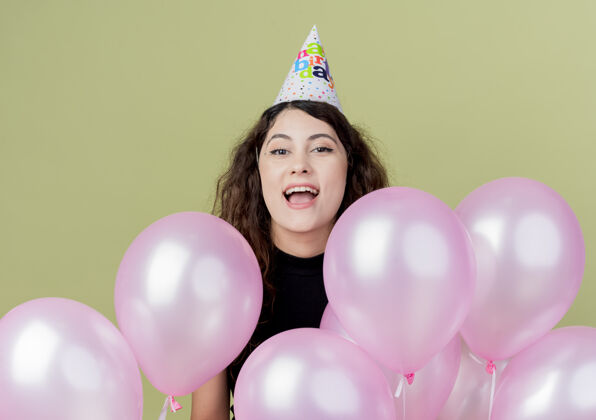 帽子一个年轻漂亮的女人 卷发 戴着节日帽 手里拿着气球 站在灯火通明的墙上欢庆生日庆祝年轻女人